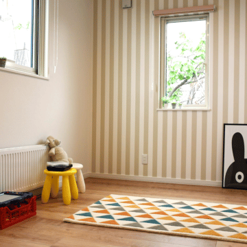 木目調の床にベージュのストライプ柄の壁紙で、北欧テイストのおもちゃやラグが置かれた洋室1です。