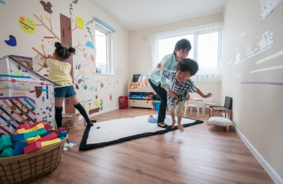 木目調の床にはおもちゃがいっぱいあり、女の子が壁一面にシールを貼って遊べんでいる。お父さんと男の子は飛行機ごっこをして遊んでいる洋室2です。