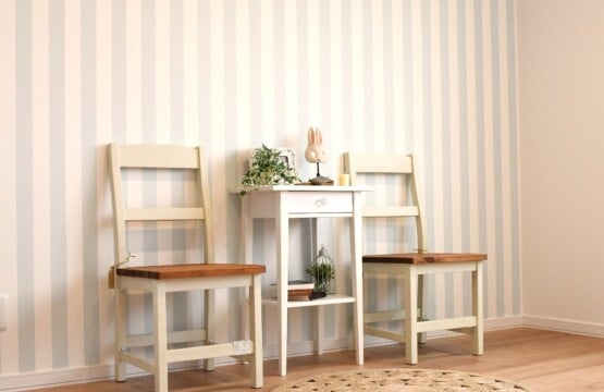 木目調の床に薄いブルーのストライプ柄の壁紙、壁にはナチュラルテイストの木の椅子とサイドテーブルが置かれた洋室2です。
