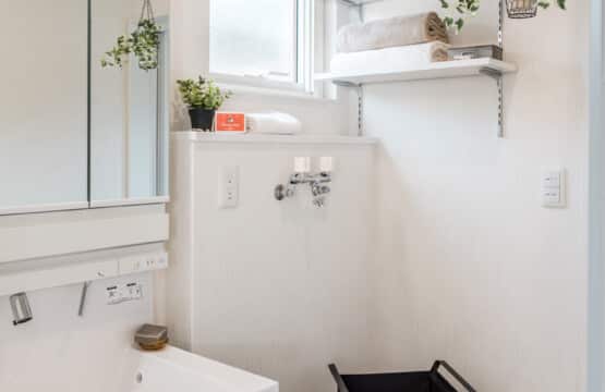 収納の扉が鏡になった洗面台の側面の収納部分はダークブラウンになっており、洗面台のすぐ右側は洗濯機を置くスペースになっています。洗濯機を置くスペースの上部には小さなスクエア窓が付いており、右側の壁には白い可動棚が付いていてタオルを収納しています。洗面化粧室の上には洗濯物を干せるポールが付いている、ホワイトナチュラルの洗面化粧室です。