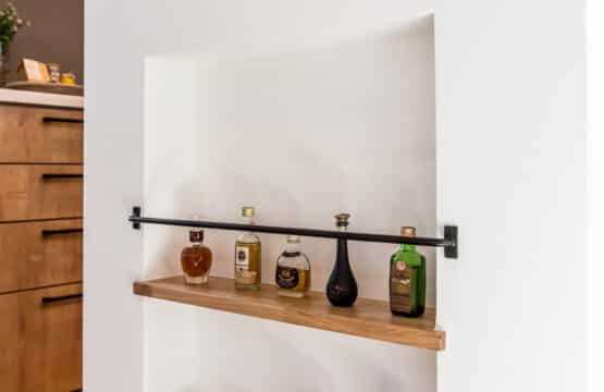 キッチンの白い立ち上がり壁の側面には2段のボトルニッチがあり、複数の瓶が飾られています。ボトルニッチの2段の棚板は、立ち上がり壁の天板と同じ木材で、黒いアイアンのストッパーが付いており、ボトルが簡単に落ちないようになっています。ボトルニッチのある壁の奥には木製のカップボードが見えています。