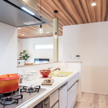 キッチン全体は白く、キッチンの立ち上がり壁の上部にはソケットのペンダントライトが横並びに２つ吊り下げられています。ガスコンロにはル・クルーゼのようなオレンジの鍋が飾られています。キッチンの後ろ側にはダークブラウンの収納棚が見えています。白いクロスに白系の木材の床、天井はブラウン系の木材のペントハウスのキッチンです。