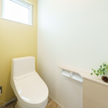テラコッタ調の床、右側壁がレモンイエロー色、左側の壁は白色で2連の紙巻き器が付いたトイレカウンター収納が設置されているおトイレです。
